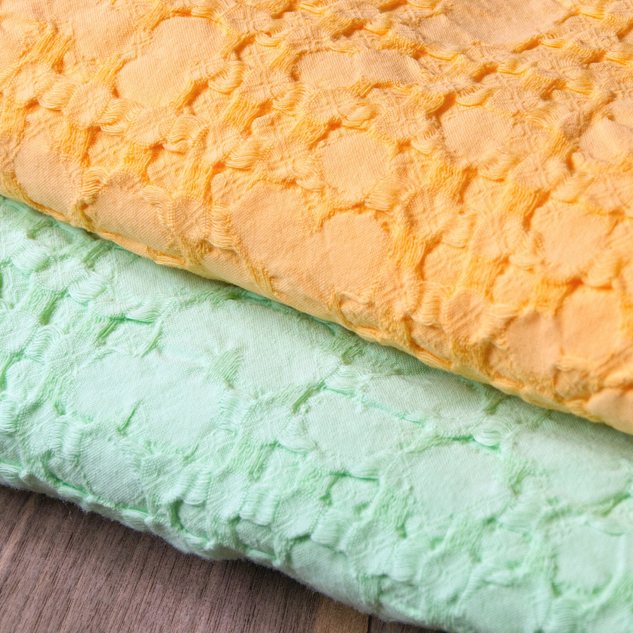 Close-up of textured fabric of two  wearable throw blankets, one mango and one pistachio, on a wooden surface.