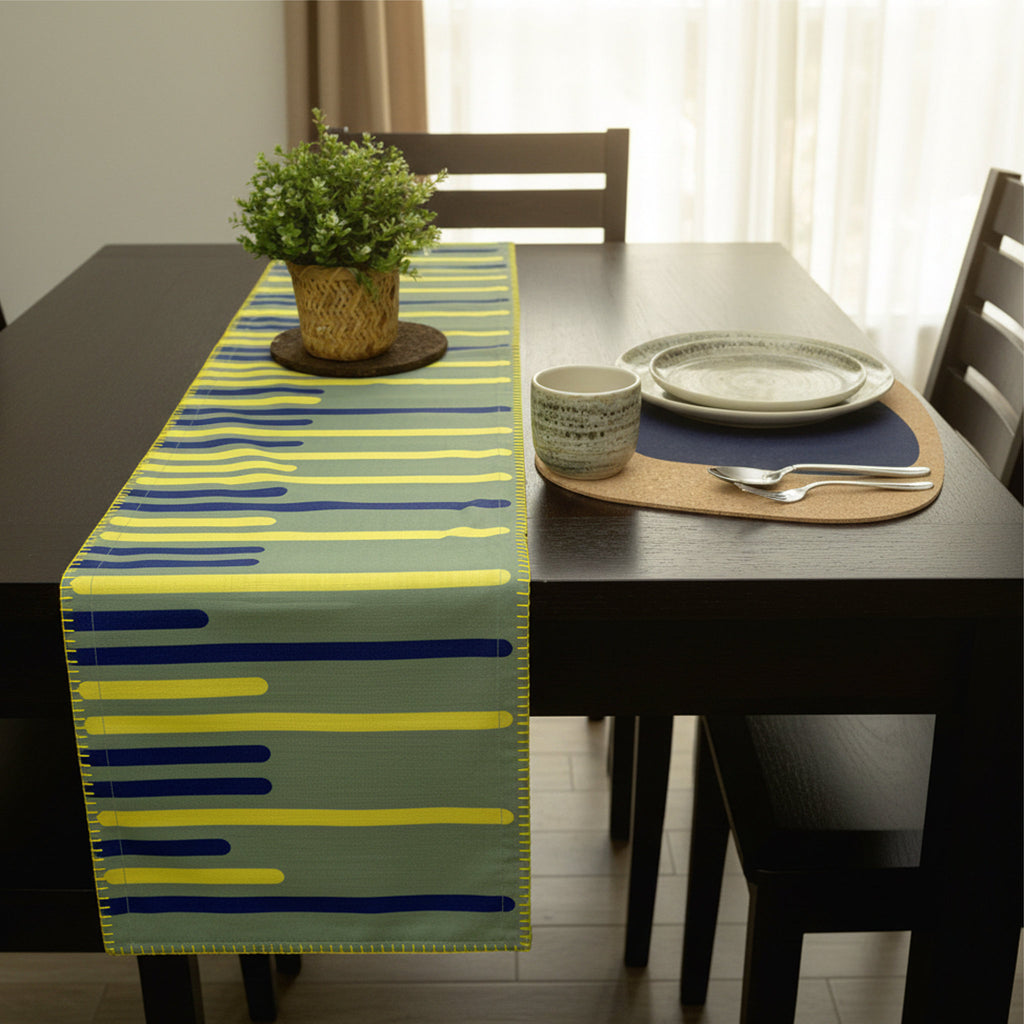 A table runner with a modern abstract stripe pattern in yellow, blue, and green, styled on a dark wooden dining table with a potted plant and place settings from Hem And Tassel 
