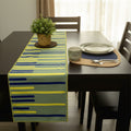 A table runner with a modern abstract stripe pattern in yellow, blue, and green, styled on a dark wooden dining table with a potted plant and place settings from Hem And Tassel 