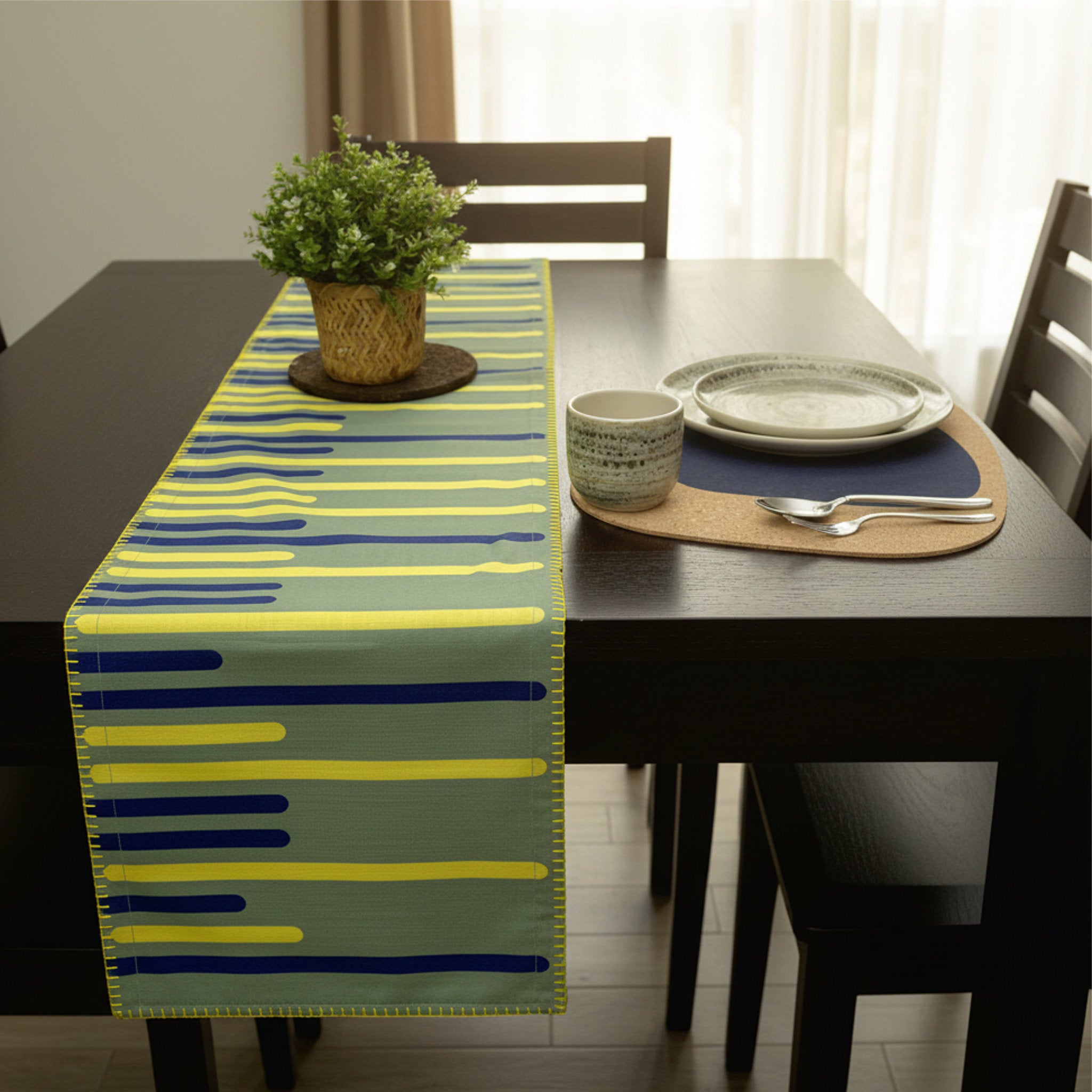 A table runner with a modern abstract stripe pattern in yellow, blue, and green, styled on a dark wooden dining table with a potted plant and place settings from Hem And Tassel 