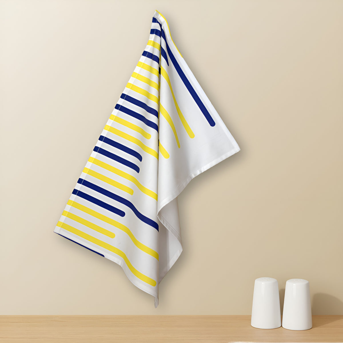 A cotton kitchen towel with yellow and navy stripes running randomly too look like piano keys, as part of the yellow piano collection, on a white background, hanging against a beige wall above a wooden surface with salt and pepper shakers below from Hem And Tassel