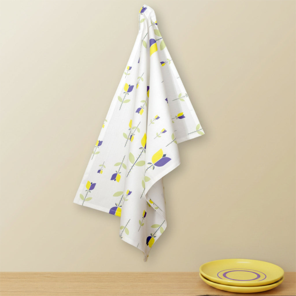 A white kitchen towel with a yellow and purple floral pattern hangs from a hook on a beige wall above a wooden surface with yellow plates from Hem And Tassel
