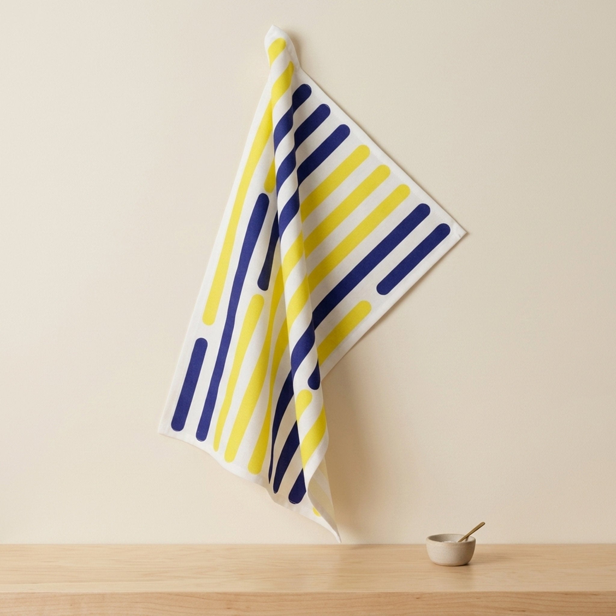 A white cotton kitchen towel with a navy and yellow intermittent stripes of the yellow piano collection hangs from a hook on a beige wall above a wooden table with a small ceramic bowl and spoon from Hem And Tassel