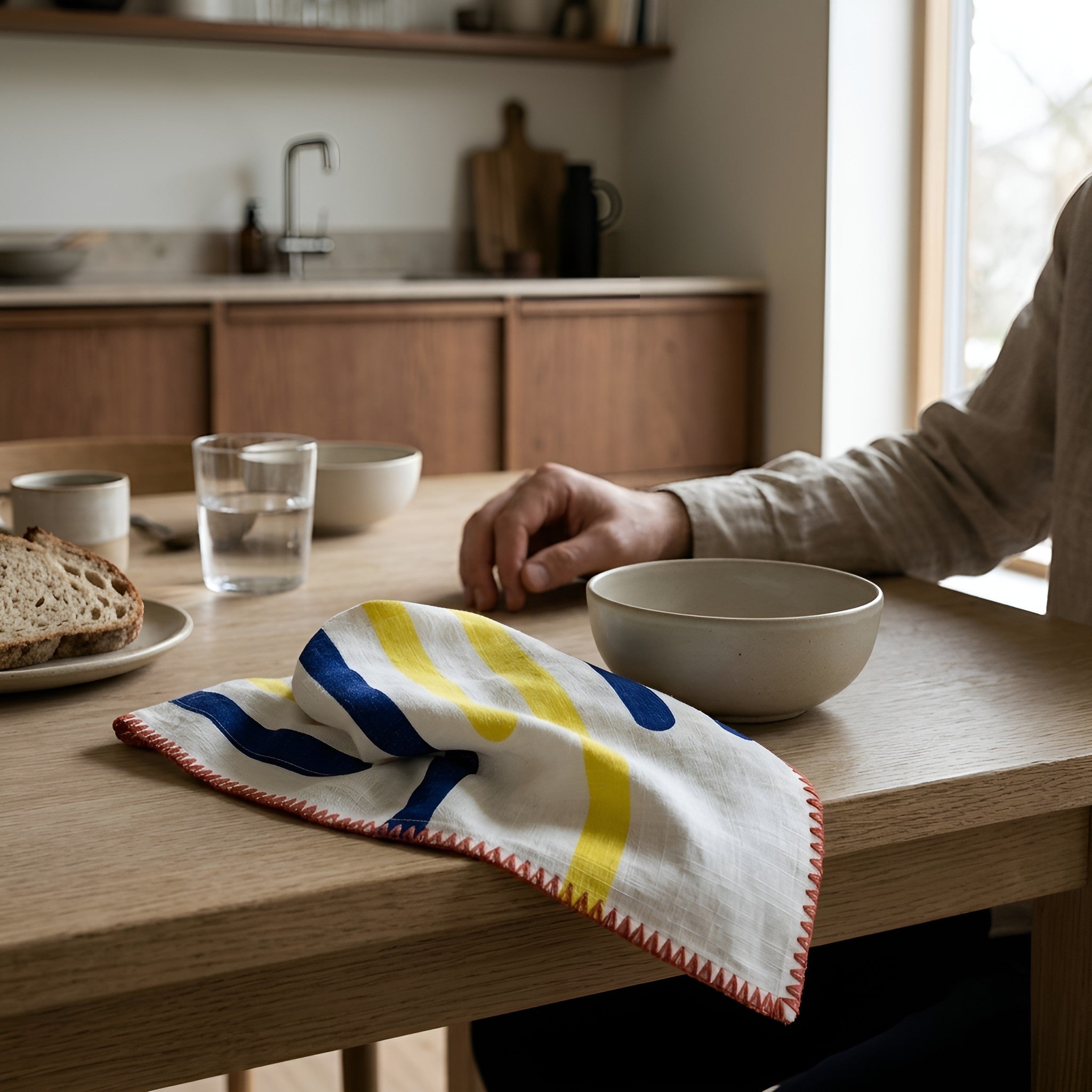 A yellow piano dinner napkin with blue and yellow stripes and red zigzag edges, styled on a wooden dining table in a bright kitchen setting with a person's hand resting nearby from Hem And Tassel 