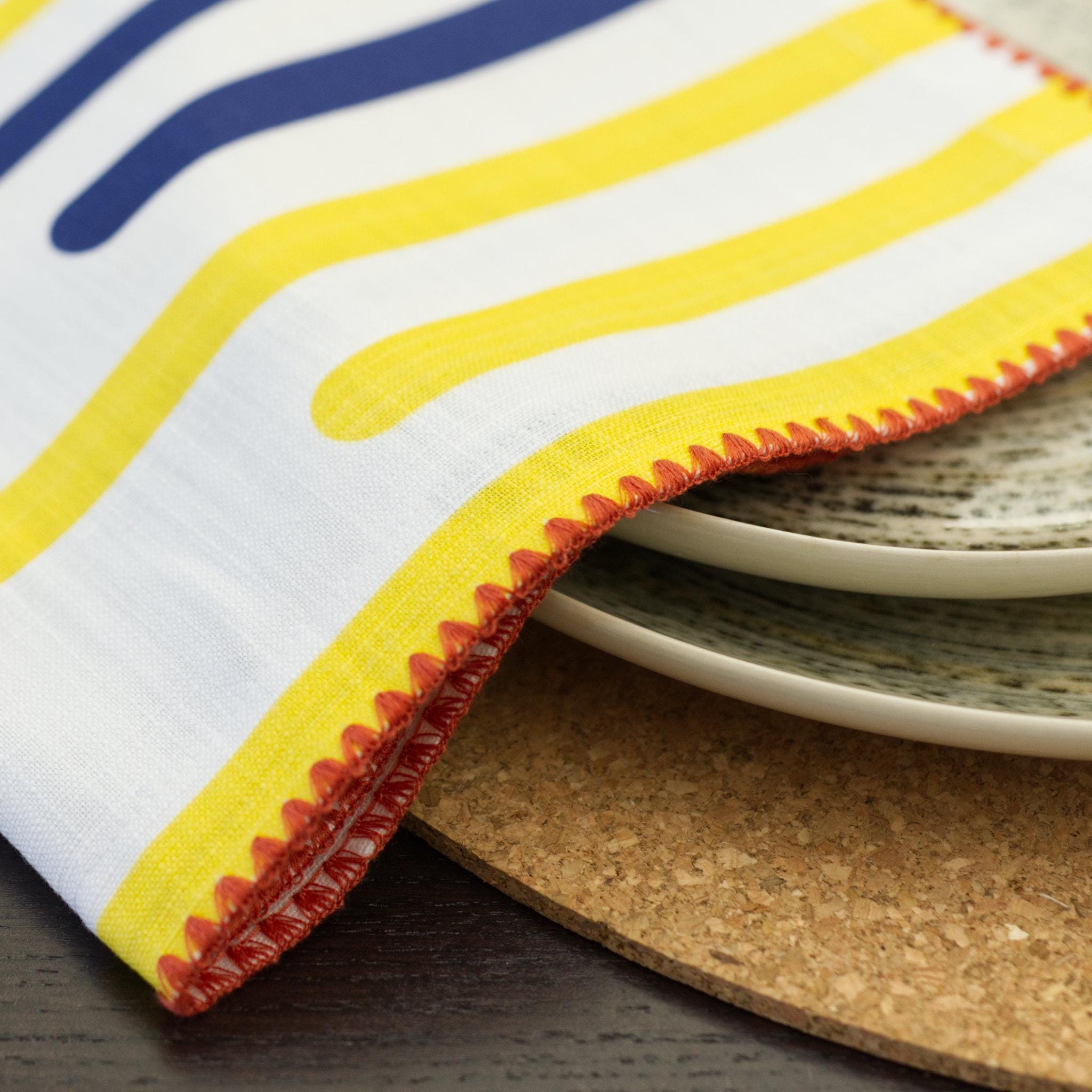 closeup of decorative detail on yellow piano dinner napkin in pink stitching, with plates on a cork placemat, from Hem And Tassel