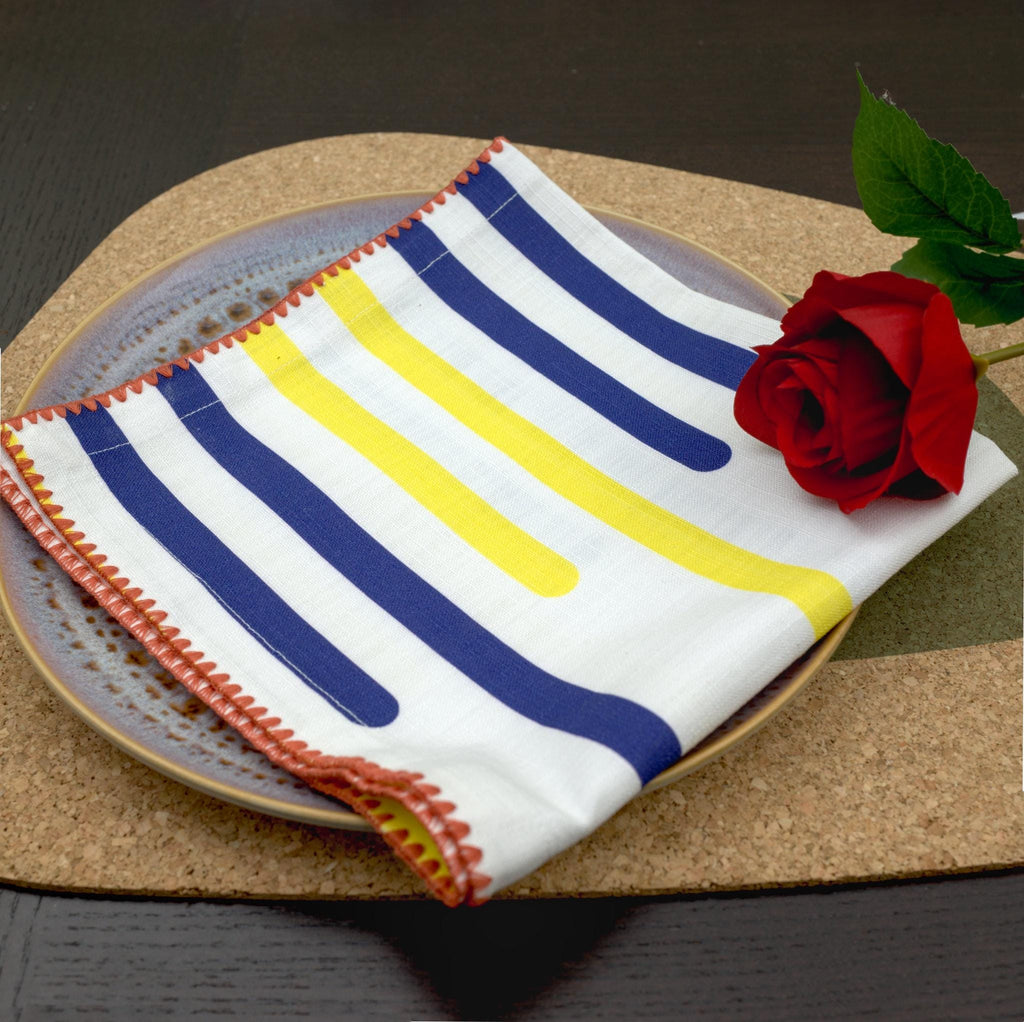 closeup of decorative detail on yellow piano dinner napkin in pink stitching, with a rose on top sitting on. plate  on a cork placemat, from Hem And Tassel