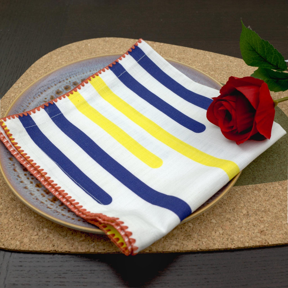 closeup of decorative detail on yellow piano dinner napkin in pink stitching, with a rose on top sitting on. plate  on a cork placemat, from Hem And Tassel