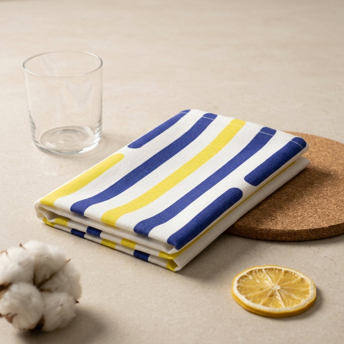 A folded striped cotton kitchen towel  with navy blue and yellow vertical stripes on a white base, styled on a beige surface with a glass, cork coaster, cotton boll, and dried lemon slice from Hem And Tassel