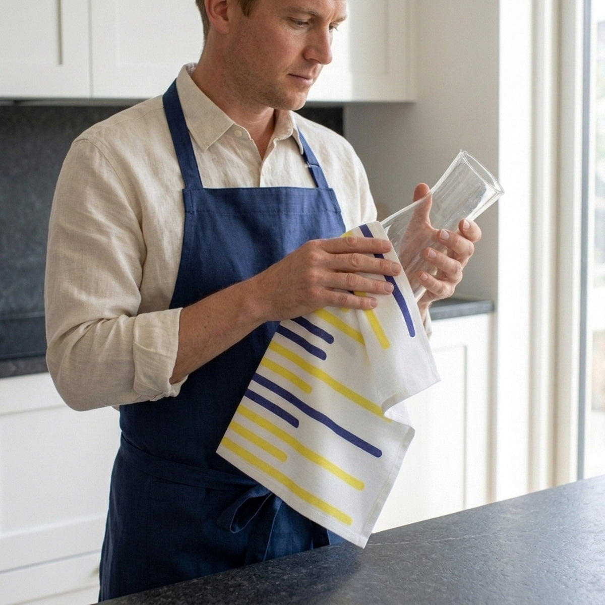 A man in a kitchen wearing a navy blue apron over a light beige shirt, holding a clear glass tumbler and a white kitchen towel with yellow and navy blue stripe patterns from Hem And Tassel
