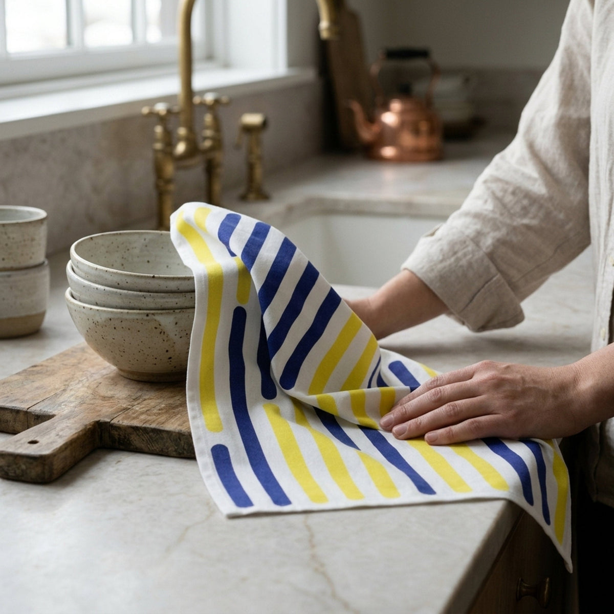 A striped kitchen towel with blue, yellow, and white stripes is draped over a marble countertop in a kitchen setting, with a person's hands and arms visible holding and smoothing the towel from Hem And Tassel