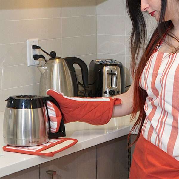 girl in kitchen wearing everyday classic apron in red, showing her wearing a decade classics oven mitt in red and a pot holder acting as a trivet