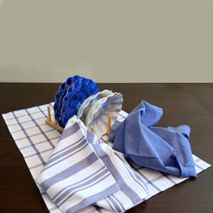 Three large 100% cotton kitchen towels in flat weave shown on a dining table, of decade classic style, in solid blue, checks and stripes