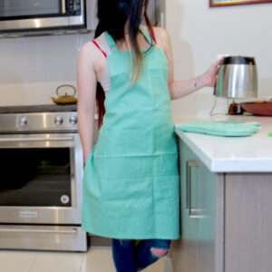 Girl wearing, pro-grade, heavy duty, drill cotton cooking and baking Apron, in jade green with divided pocket and with adjustable neck straps. With matching oven mitt and pot holder