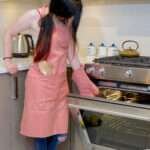 Girl wearing, pro-grade, heavy duty, drill cotton cooking and baking Apron, in rose pink with divided pocket and with adjustable neck straps. With matching oven mitt and pot holder