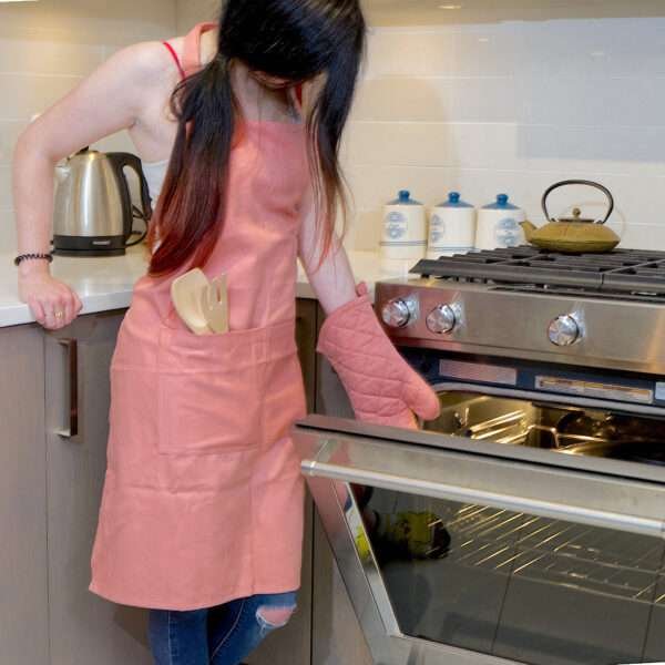 Girl wearing, pro-grade, heavy duty, drill cotton cooking and baking Apron, in rose pink with divided pocket and with adjustable neck straps. With matching oven mitt and pot holder