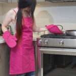 Girl at grill wearing, pro-grade, heavy duty, drill cotton cooking and baking Apron, in fuchsia with divided pocket and with adjustable neck straps. With matching oven mitt and pot holder