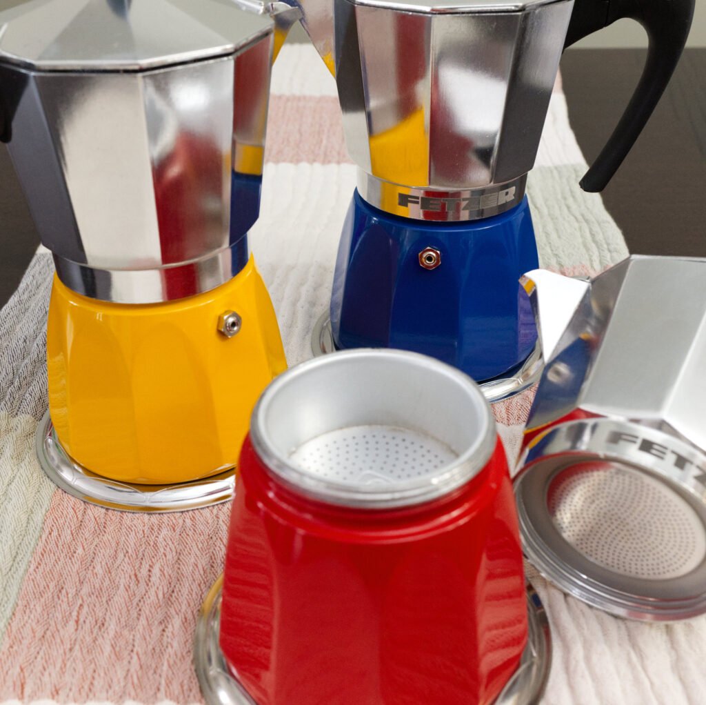 Induction moka pots in red, yellow and blue colors all sitting on a table runner