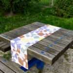 Table runner of flock and flutter design in clay on picnic table in park from Hem And Tassel