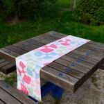 Table runner of flock and flutter design on picnic table in park from Hem And Tassel