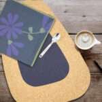 Shy Dragonfly-dinner napkin in Oil+Kidney Bean cork placemat in Blue on a table with coffee and a spoon from hem and tassel